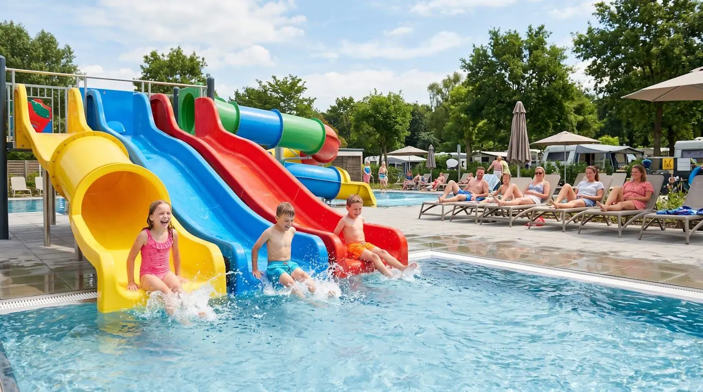 Des enfants rient et jouent dans une piscine de camping avec toboggans colorés, surveillés par leurs parents assis sur des transats en arrière-plan sous un soleil d'été